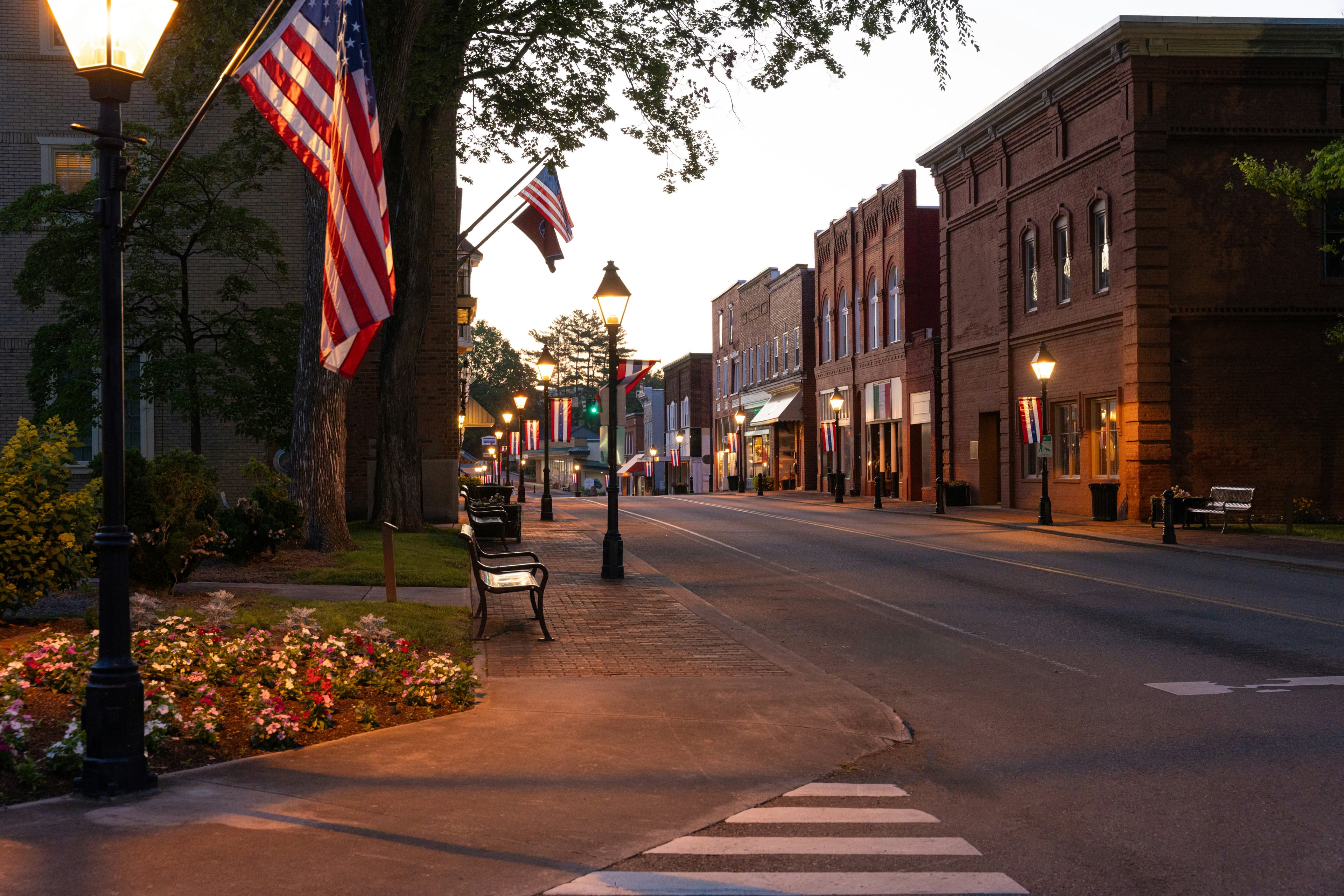 A small town main street at dusk lined with American flags, brick buildings, and street lamps